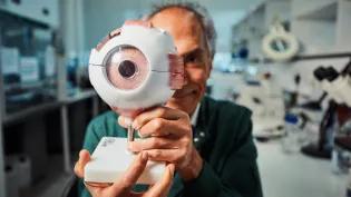 Researcher Baljean Dhillon looking happy holding a large 3D model of an eyeball. He is wearing a green lab coat and is standing at a workbench in a laboratory