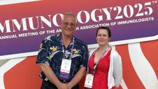 Adriano Rossi (in hawaiian shirt) and Jennifer Cartwright (in red dress) smiling in front of an 'Immunology2025' poster. 