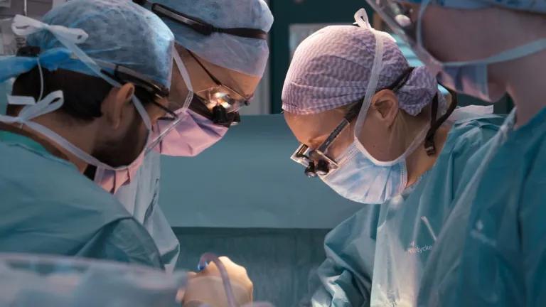 Two surgeons with face masks, glasses, surgical overalls and hair coverings looking down as they operate