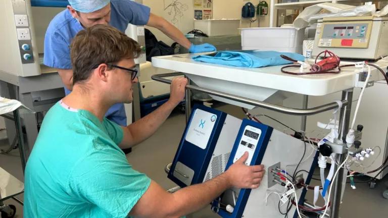 clinician bending down in green scrubs and pressing buttons on a perfusion machine, roughly 1m by 0.5m