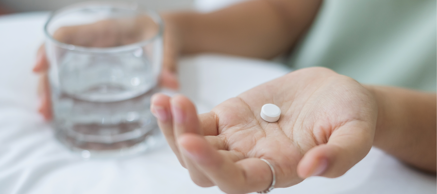 Close-up photo of a woman's Hands. One holds a small white pill and the other holds a small glass of water.