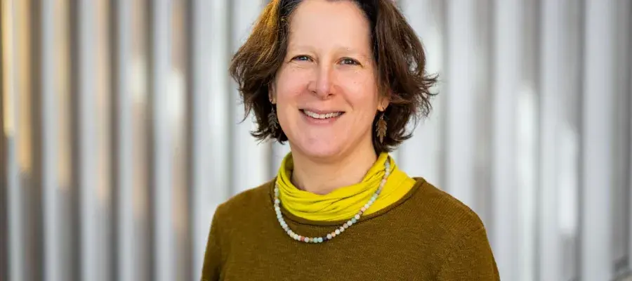A woman with short brown hair smiles at the camera. She is wearing a brown knit jumper and a yellow turtleneck.