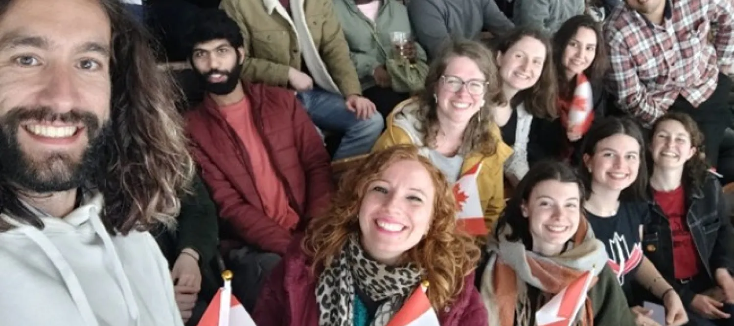 a group of people sat in the stands of an ice hockey rink, many are holding small Canadian flags