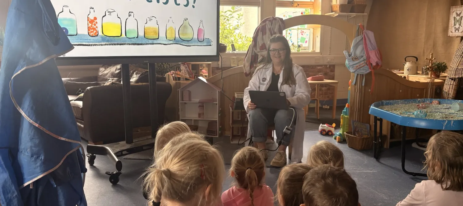 A researcher sitting on a low chair in front of a classroom of children