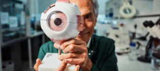 Researcher Baljean Dhillon looking happy holding a large 3D model of an eyeball. He is wearing a green lab coat and is standing at a workbench in a laboratory