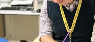 Neil Ryan is standing in an office with a computer in the background. He is looking and pointing at an image in a textbook called "Modern Pathology". He is wearing a striped collared shirt and a blue sleevless jumper