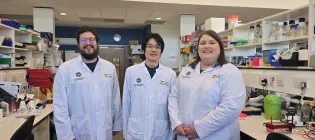 Three researchers stand in a lab wearing lab coats. They are all smiling at the camera