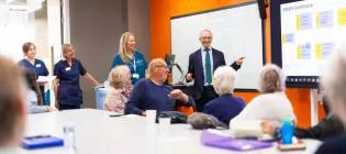 Boardroom with Adam Hill presenting to a room, and patients and nurses are there