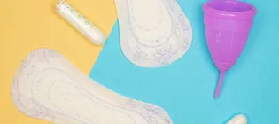 A selection of period pads, tampons and a purple moon cup on a yellow and blue background