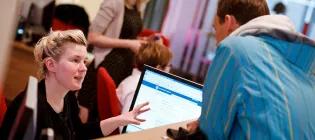 UoE staff member at a desk talking to a student and gesturing to a screen