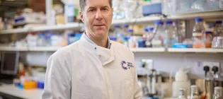 Steve Pollard in a lab coat standing in front of a lab bench with lots of bottles on the shelves behind him