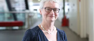 Professor Anna Williams has short white hair and is wearing glasses. She is stood in a hallway in front of a staircase