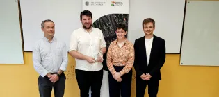 Andrew McBryde, Dan Edwards, Becca Belmonte, and Marcin Kedziera stand in front of a yellow wall and a pop-up banner. Dan is holding a glass trophy