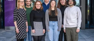 The Gibson group of researchers. 7 researchers stand in a row in front of a glass building