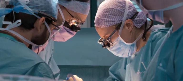 Two surgeons with face masks, glasses, surgical overalls and hair coverings looking down as they operate