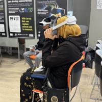 A boy and woman sit down in a hall with VR headsets and a poster behind titled FishEye Reality