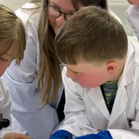 A boy looking down a microscope with another boy at his side and a voluneer helping in the background