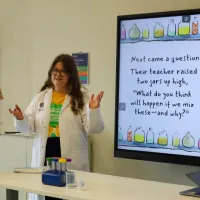 Kelly holding 2 conical flasks and Lucy with both arms up in front of a screen