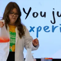 Lucy holding a giant conical flask with bright orange liquid inside that's smoking