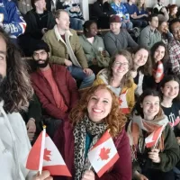 a group of people sat in the stands of an ice hockey rink, many are holding small Canadian flags