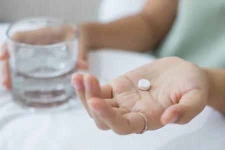 Close-up photo of a woman's Hands. One holds a small white pill and the other holds a small glass of water.