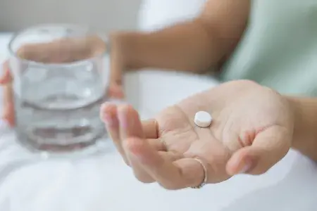 Close-up photo of a woman's Hands. One holds a small white pill and the other holds a small glass of water.