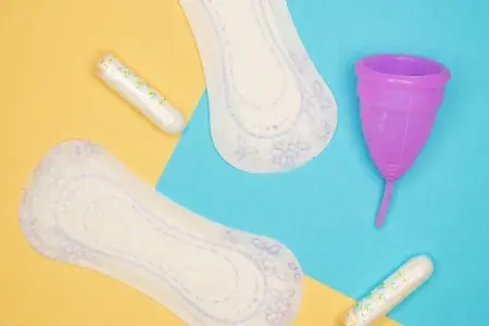 A selection of period pads, tampons and a purple moon cup on a yellow and blue background