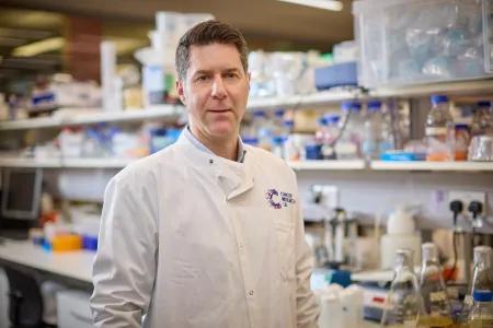 Steve Pollard in a lab coat standing in front of a lab bench with lots of bottles on the shelves behind him