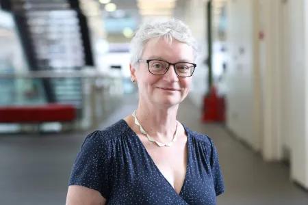 Professor Anna Williams has short white hair and is wearing glasses. She is stood in a hallway in front of a staircase
