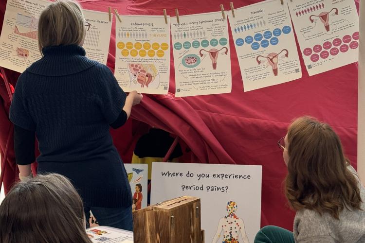 Three women of mixed ages reading informative posters on the uterus in front of a large red tent.