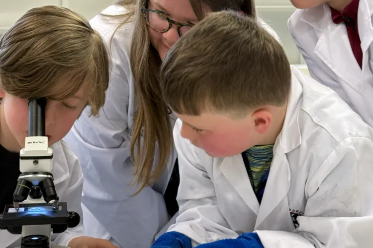 A boy looking down a microscope with another boy at his side and a voluneer helping in the background