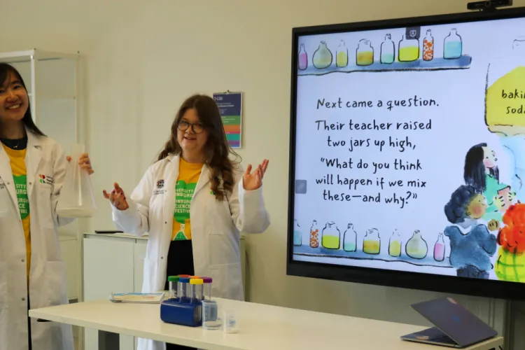 Kelly holding 2 conical flasks and Lucy with both arms up in front of a screen