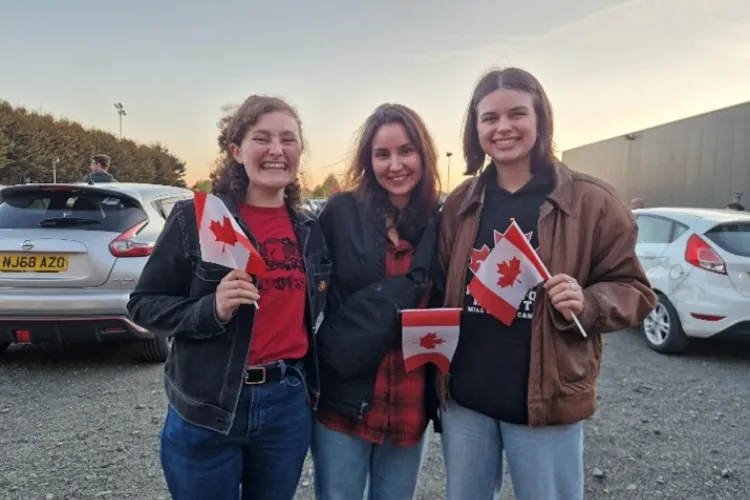Three people standing outside in a car park, all holding small Canadian flags