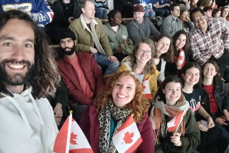a group of people sat in the stands of an ice hockey rink, many are holding small Canadian flags