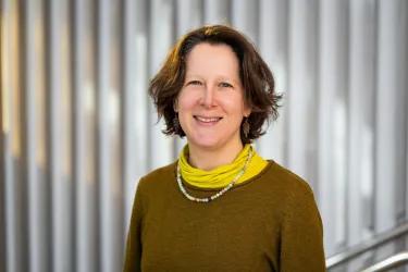 A woman with short brown hair smiles at the camera. She is wearing a brown knit jumper and a yellow turtleneck.