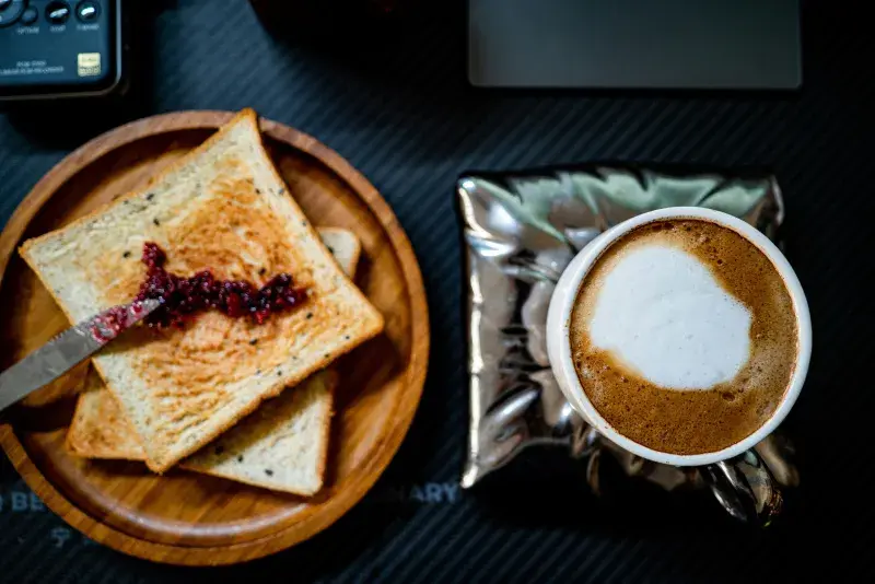 A top-down view of two pieces of toast with jam on a wooden plate, and a cup of milky coffee