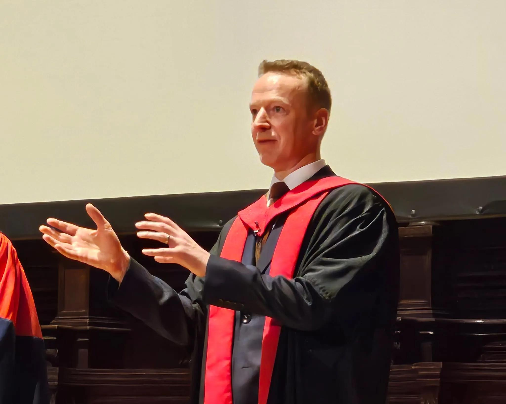 Prof Baillie in his gown, talking with his hands up