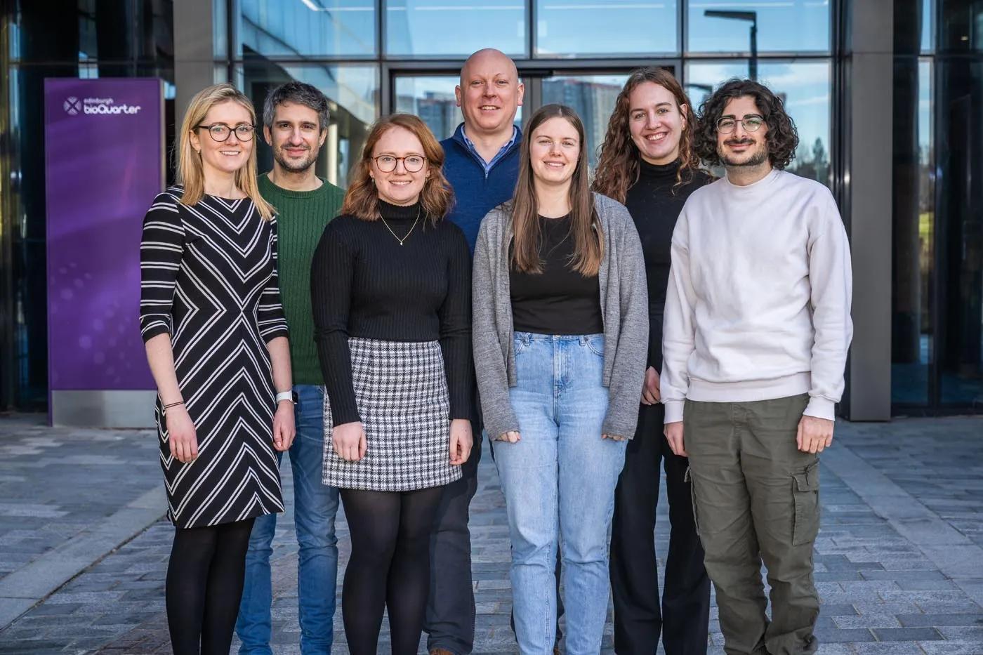 The Gibson group of researchers. 7 researchers stand in a row in front of a glass building