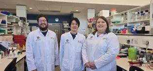 Three researchers stand in a lab wearing lab coats. They are all smiling at the camera