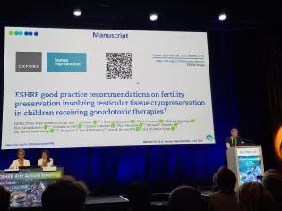 Professorr Rod Mitchell is speaking to an audience from behind a lectern with his name on it. Behind him to the left are two women at a moderator's table, and his slides are very large behind him.