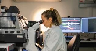 Researcher looks down microscope in a dimmed room with a lit computer screen in the background