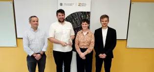 Andrew McBryde, Dan Edwards, Becca Belmonte, and Marcin Kedziera stand in front of a yellow wall and a pop-up banner. Dan is holding a glass trophy