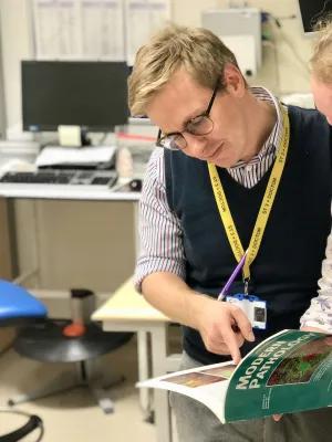 Neil Ryan is standing in an office with a computer in the background. He is looking and pointing at an image in a textbook called "Modern Pathology". He is wearing a striped collared shirt and a blue sleevless jumper