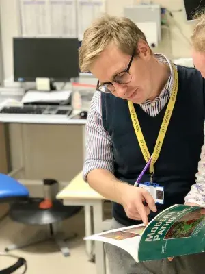 Neil Ryan is standing in an office with a computer in the background. He is looking and pointing at an image in a textbook called "Modern Pathology". He is wearing a striped collared shirt and a blue sleevless jumper