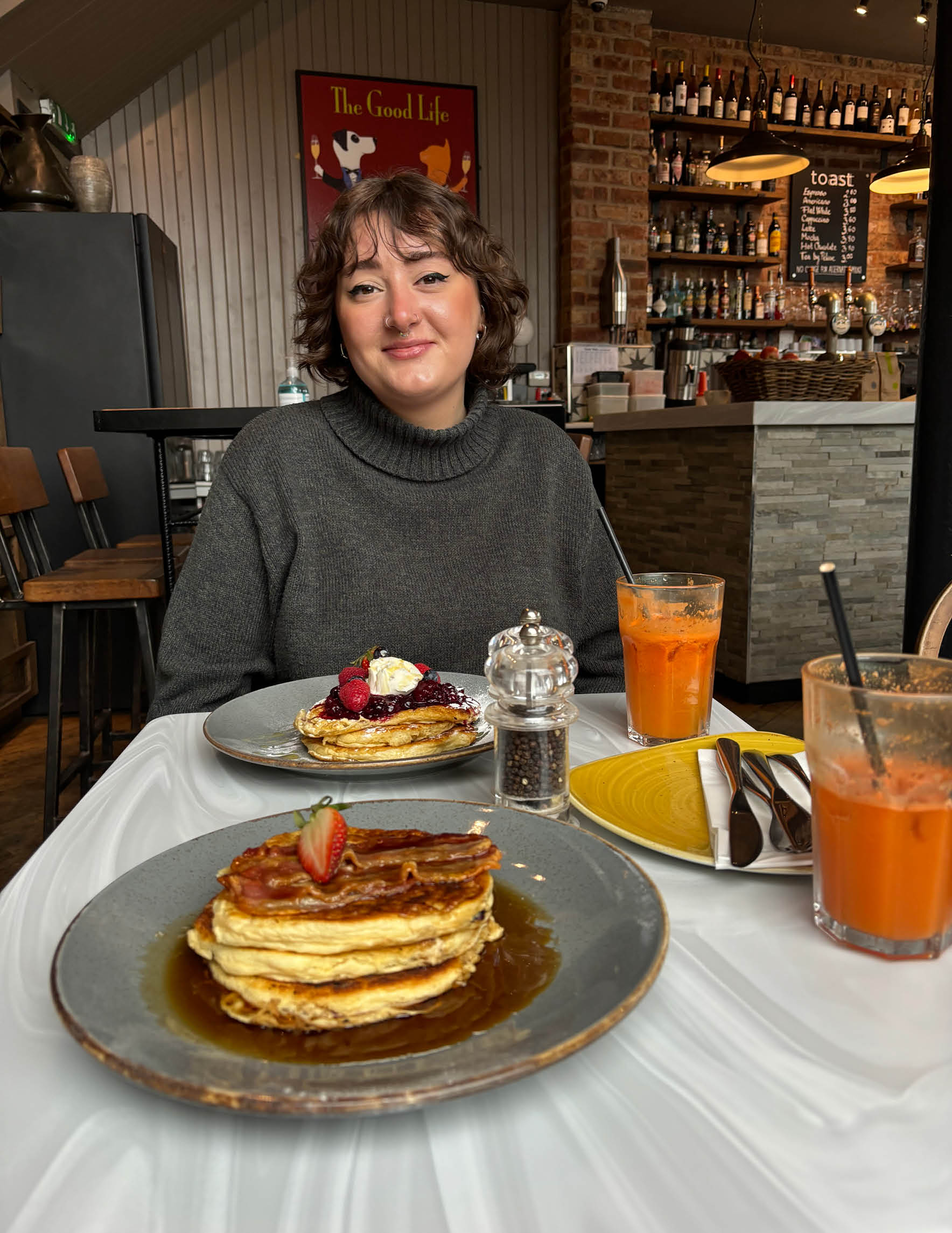 A woman with dark hair and a dark top sits at a table in front of plates of pancakes and glasses of juice