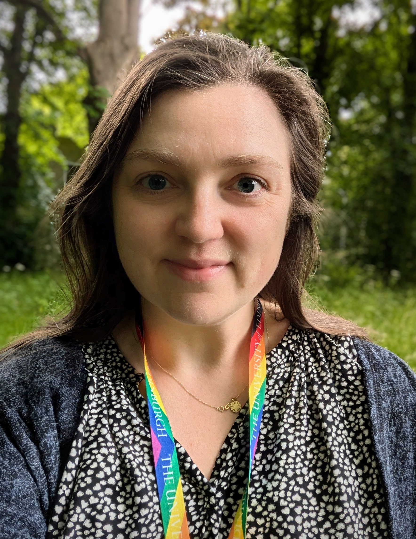 A woman wearing a rainbow lanyard and a blue top smiles at the camera in front of trees and greenery