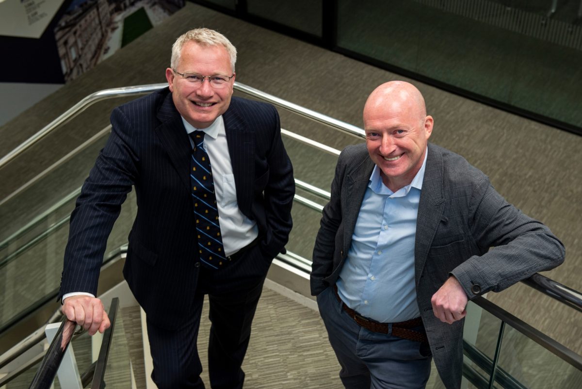 two white men in suits stand on a staircase smiling at the camera